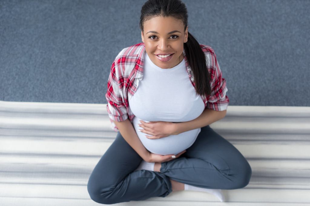 Happy Black Pregnant Woman | Columbia, MO Pregnancy | Mid-Missouri Maternal Mental Health | Postpartum Support in Missouri Smiling young black first-time mother holding her pregnant belly looking at the camera. If you are in mid-Missouri, Columbia, Ashland, Moberly, Rocheport, Holts Summit, Boonville, or surrounding areas, you may want to consider participating in this Mizzou Research Study for Maternal Mental Health.